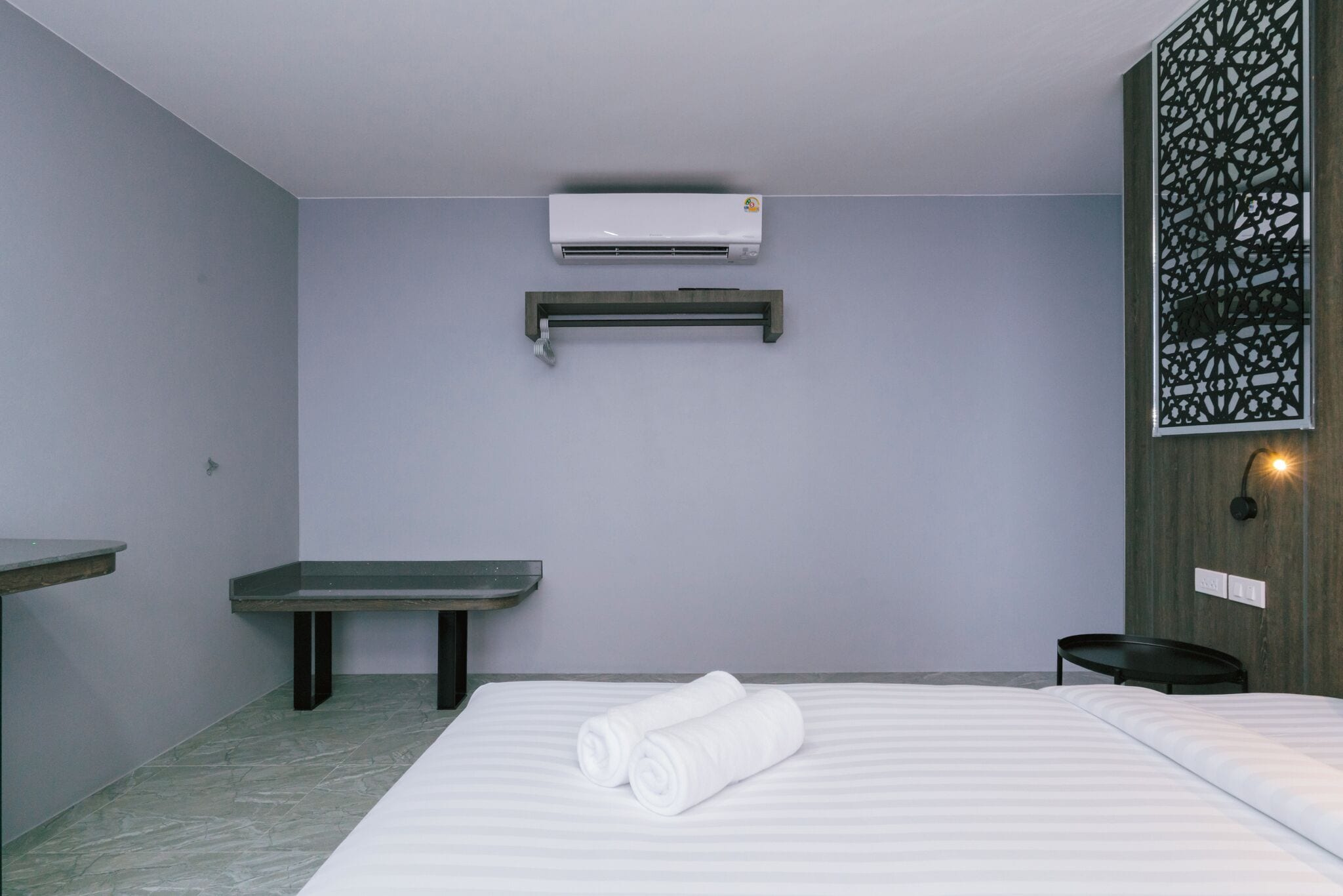 A hotel room with a bed, a bench, an air conditioning unit, and a wall-mounted light fixture.