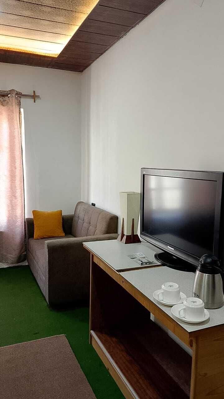 A room with a flat-screen TV, a sofa with a cushion, a coffee table with cups and a kettle, and a window with curtains.