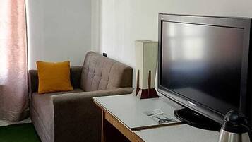 A room with a flat-screen TV, a sofa with a cushion, a coffee table with cups and a kettle, and a window with curtains.
