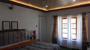 Habitación de hotel con cama, mesita de noche, un jarrón con flores, ventana con cortinas y certificados enmarcados en la pared.