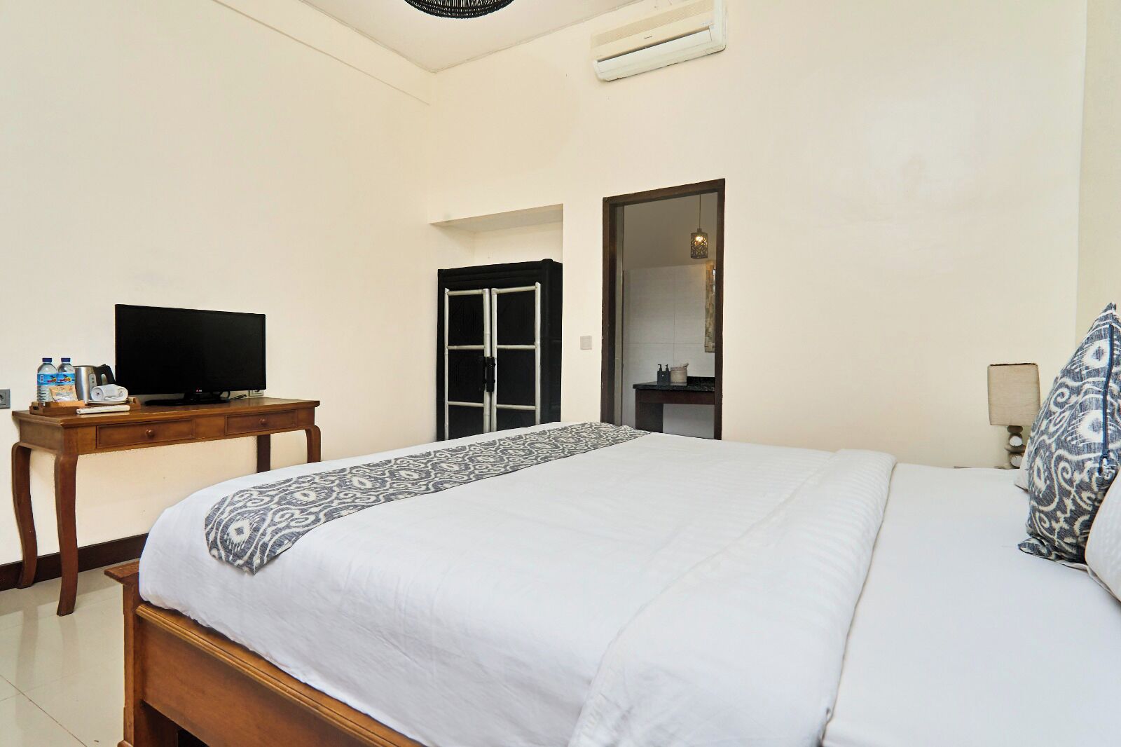 A hotel room with a bed, a television on a wooden table, a bathroom door, and a nightstand with a lamp.
