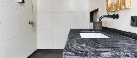 Un baño moderno con encimera de mármol, tocador de madera, un espejo grande y una ducha.