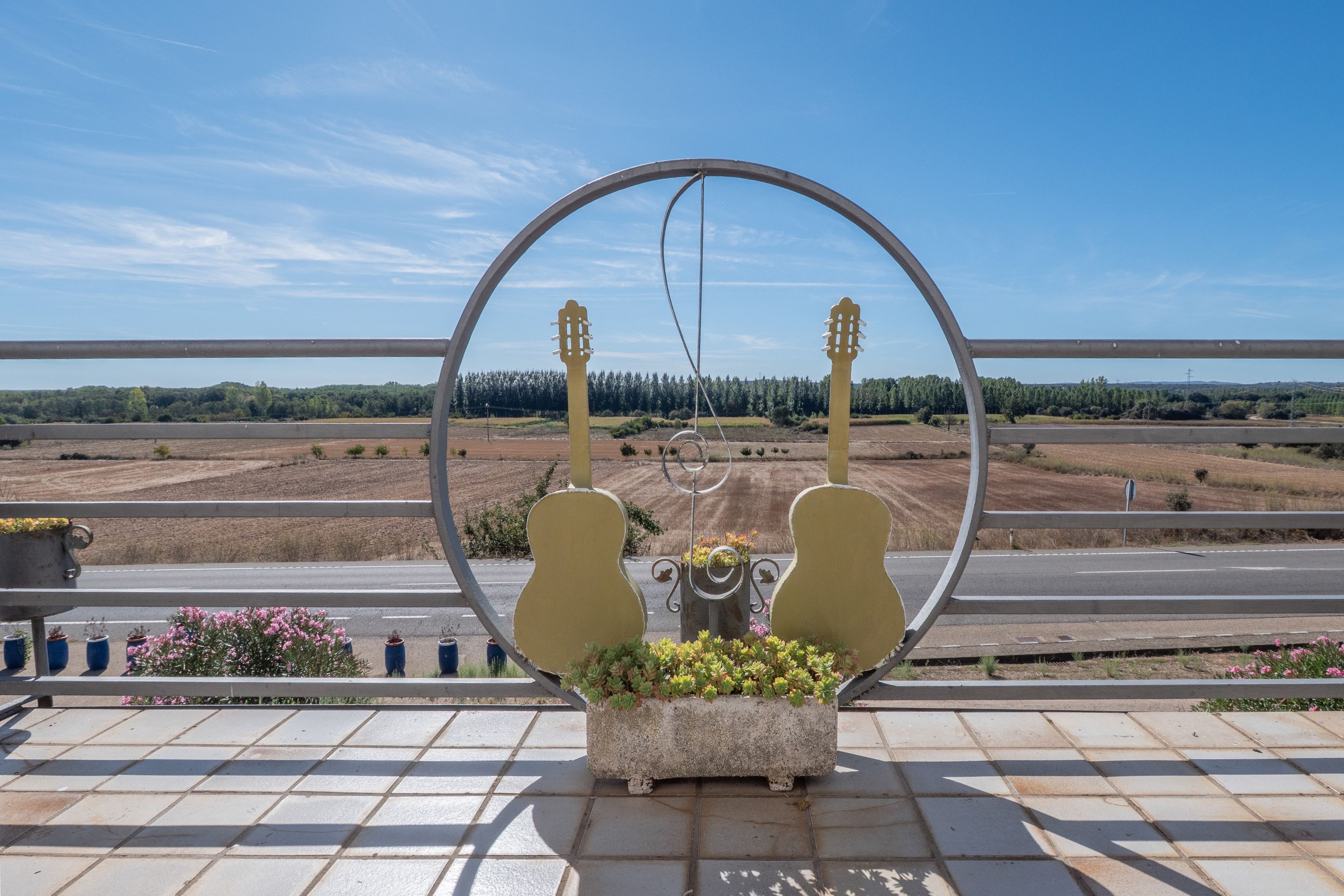 Marco metálico circular con dos esculturas con forma de guitarra y una planta en maceta al frente, sobre un fondo de carretera y árboles distantes.