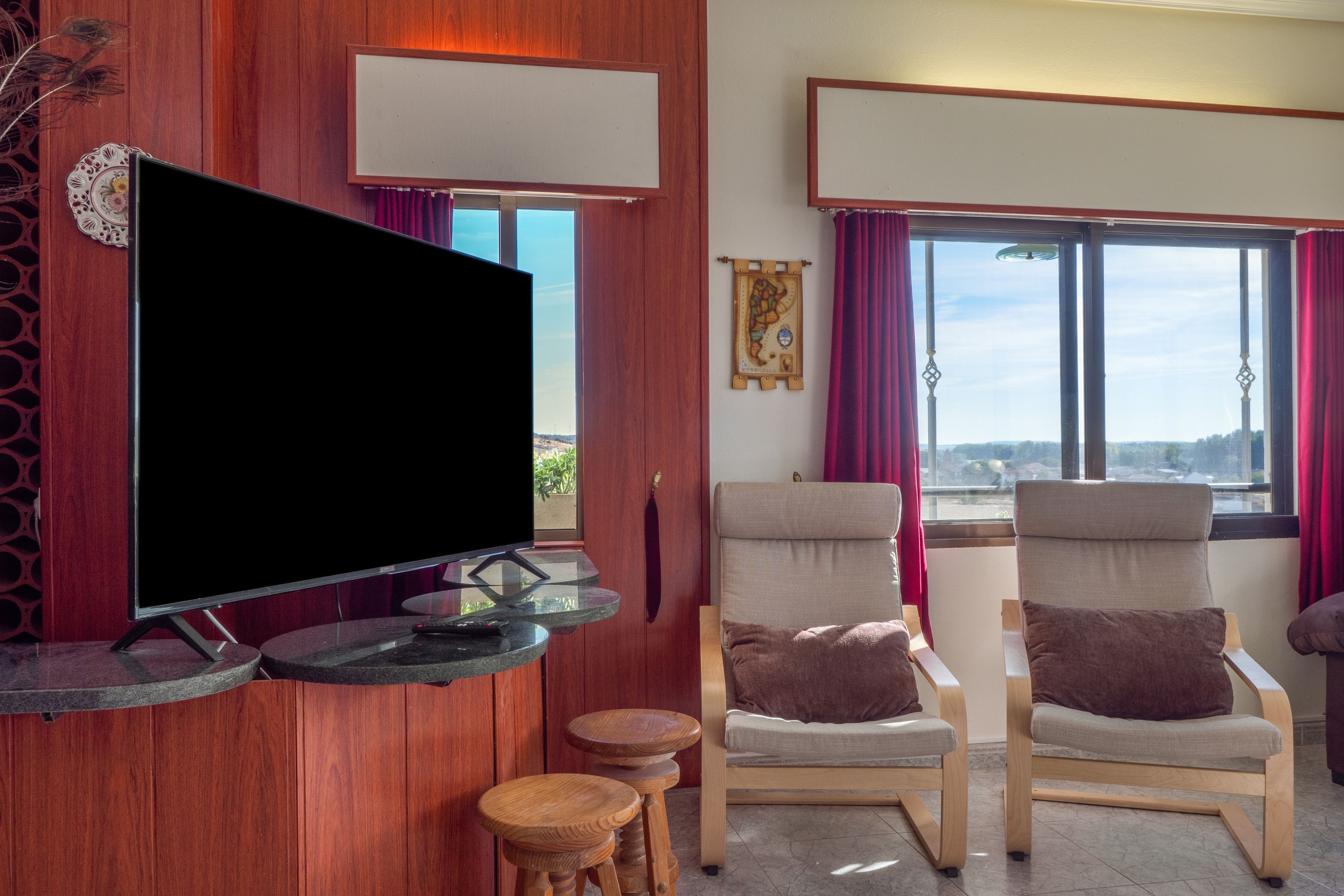 A living room with a flat-screen TV mounted on a wooden wall, two upholstered chairs with cushions, a small wooden stool, and a window with curtains.