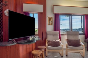 Ein Zimmer mit einem an einer Holzwand montierten Flachbildfernseher, zwei Polsterstühlen mit Kissen, einem kleinen Holzschemel und einem Fenster mit Blick ins Freie.