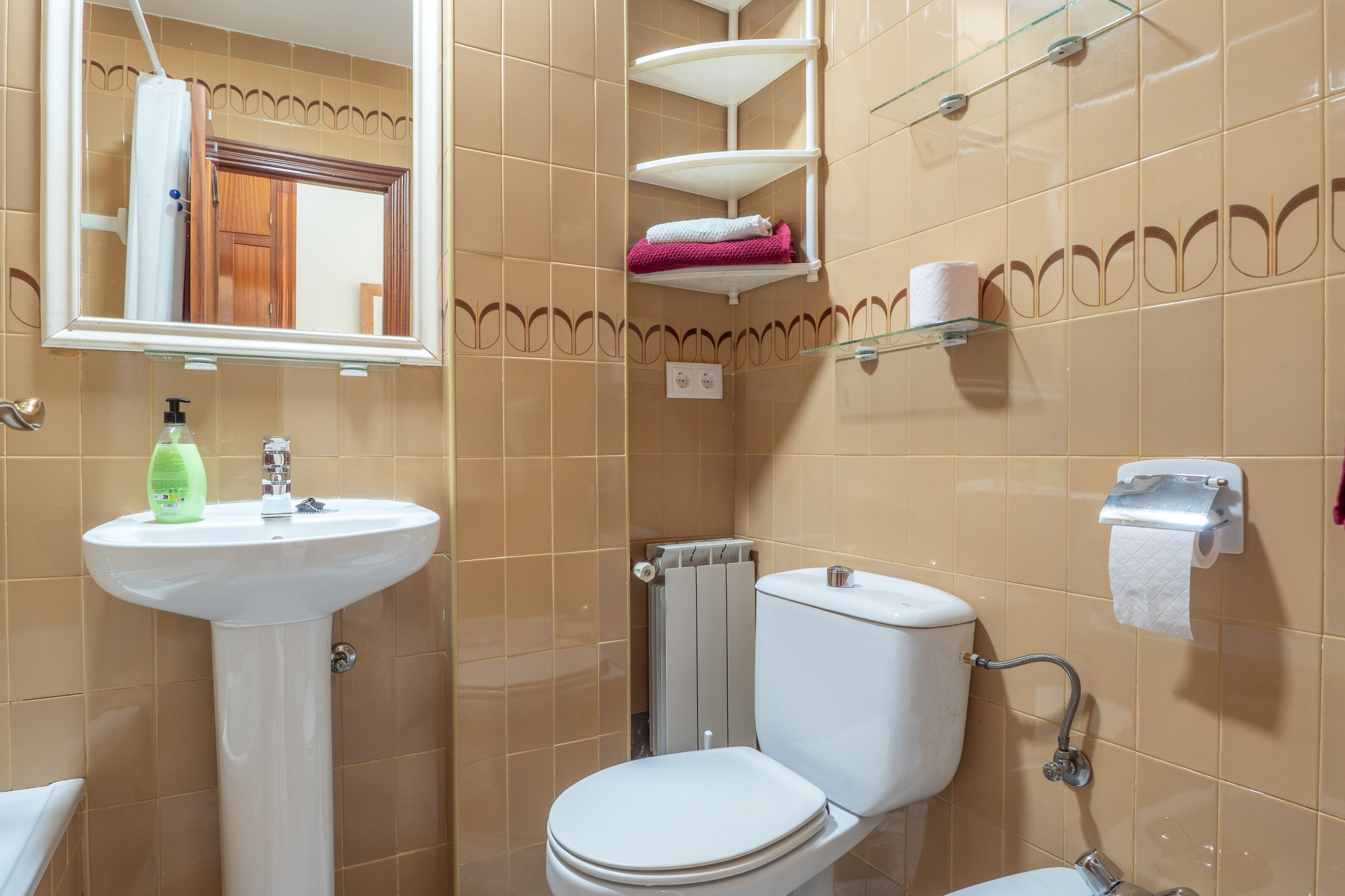 Ein Badezimmer mit weißem Waschbecken, Toilette und Dusche. Regale mit Handtüchern und ein Spiegel befinden sich über dem Waschbecken.