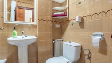 Ein Badezimmer mit weißem Waschbecken, Toilette und Dusche. Regale mit Handtüchern und ein Spiegel befinden sich über dem Waschbecken.