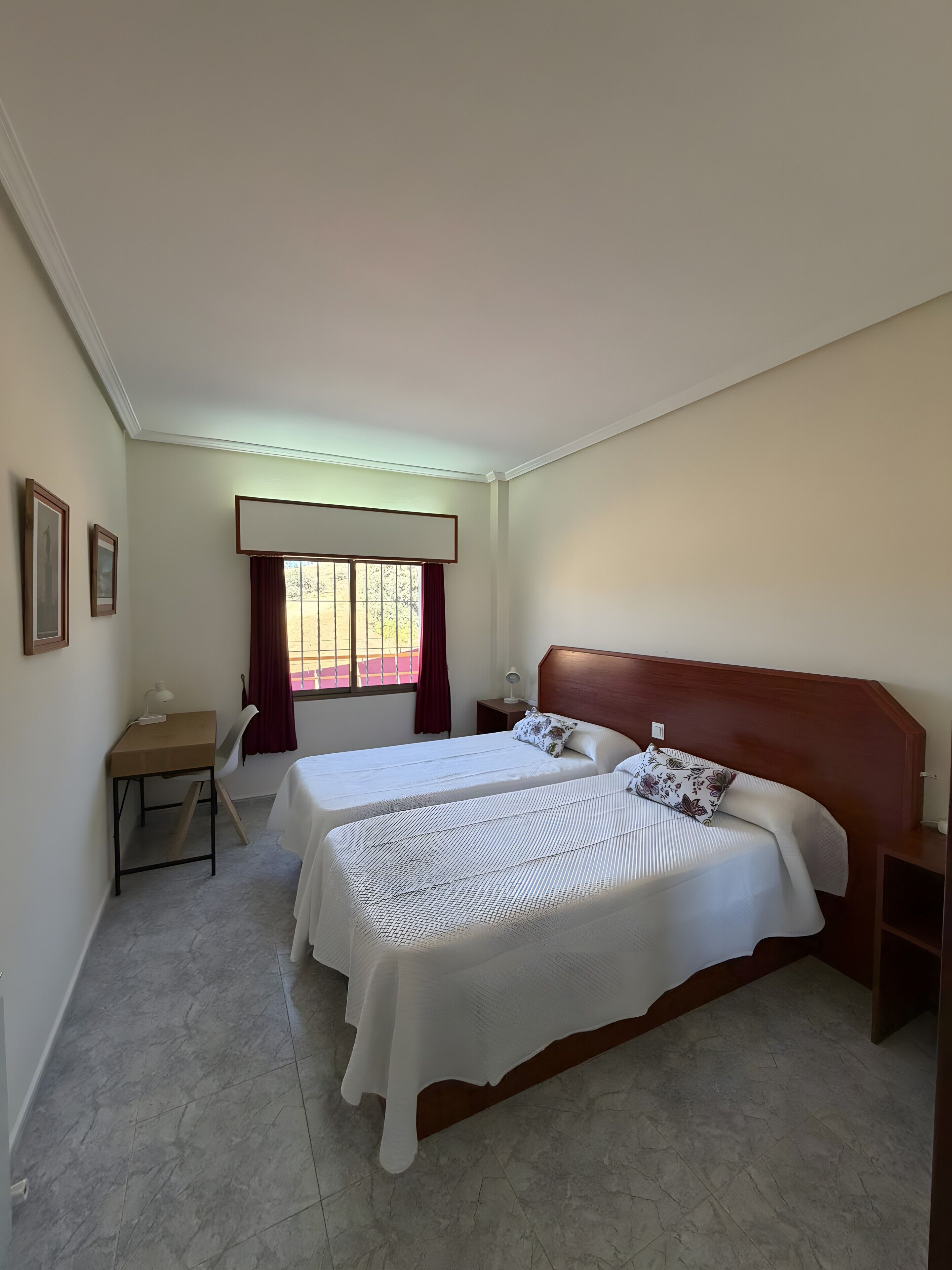 A bedroom with two beds, a desk, a window with curtains, and a nightstand.