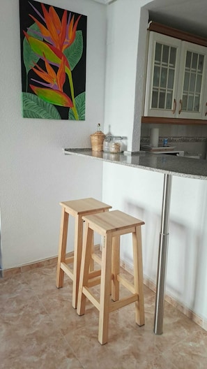 A kitchen with a white countertop, two wooden stools, a decorative plant, and a cabinet with glass doors. - Your home in Altea! 200 m from the beach / bright / 2 bedrooms / fully equipped / beautifully decorated. (Altea)