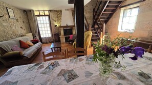 Un intérieur rustique avec un mur en pierre, un escalier en bois, un canapé, une table à manger recouverte d’une nappe ancienne et un vase rempli de fleurs.