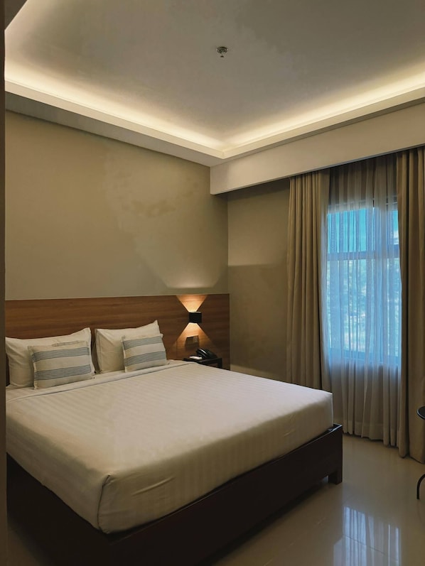 A hotel room with a large bed, a nightstand, a wall-mounted lamp, and a window with curtains.
