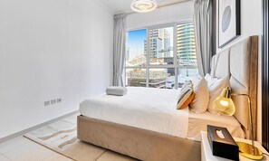 A modern bedroom with a large bed, a bedside lamp, and a view of a cityscape through large windows. - bnbme - Dunya Tower - 1BR (Dubai)