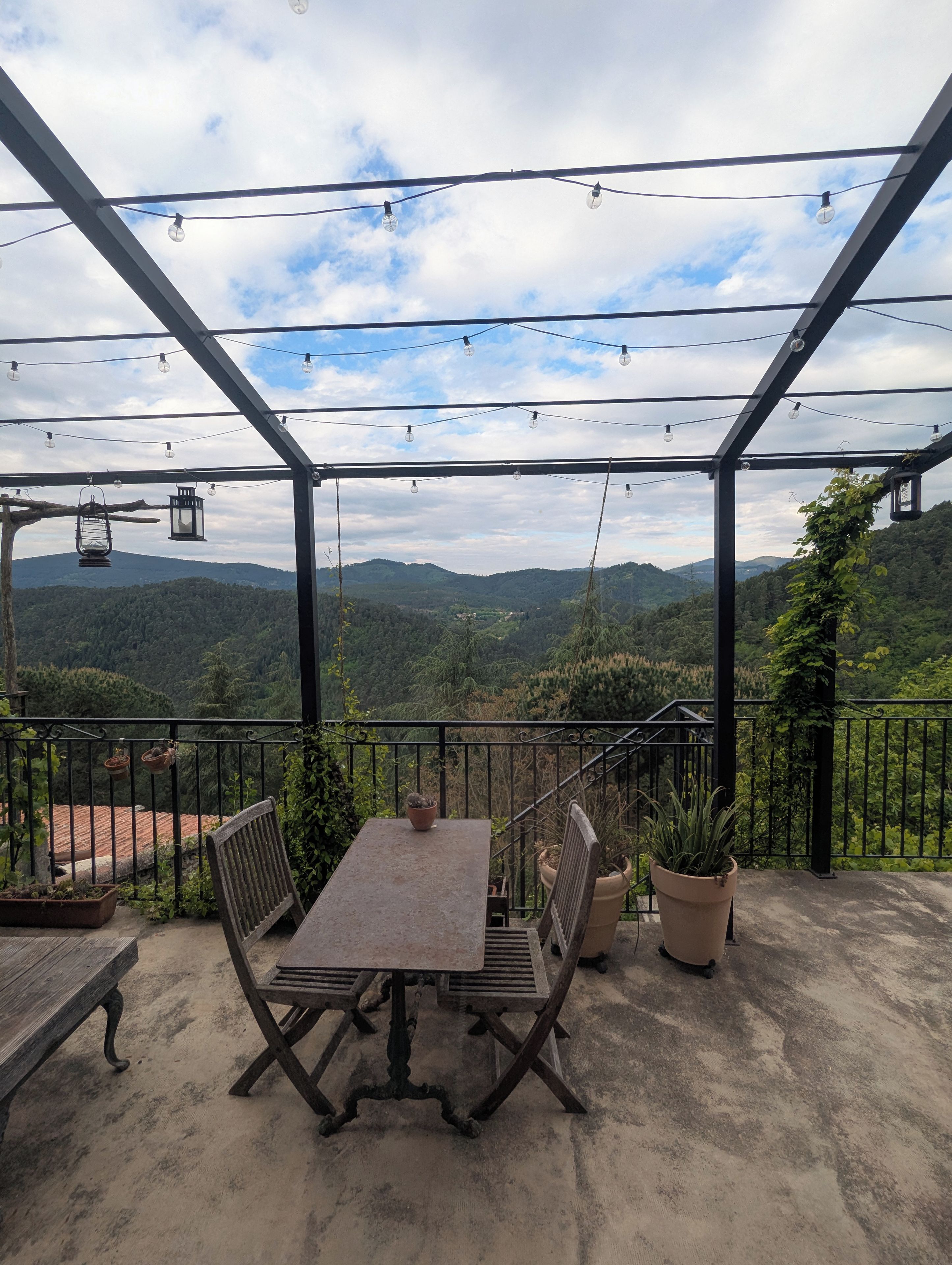 Une terrasse extérieure avec une table et des chaises, entourée de plantes en pot et offrant une vue sur un paysage montagneux.