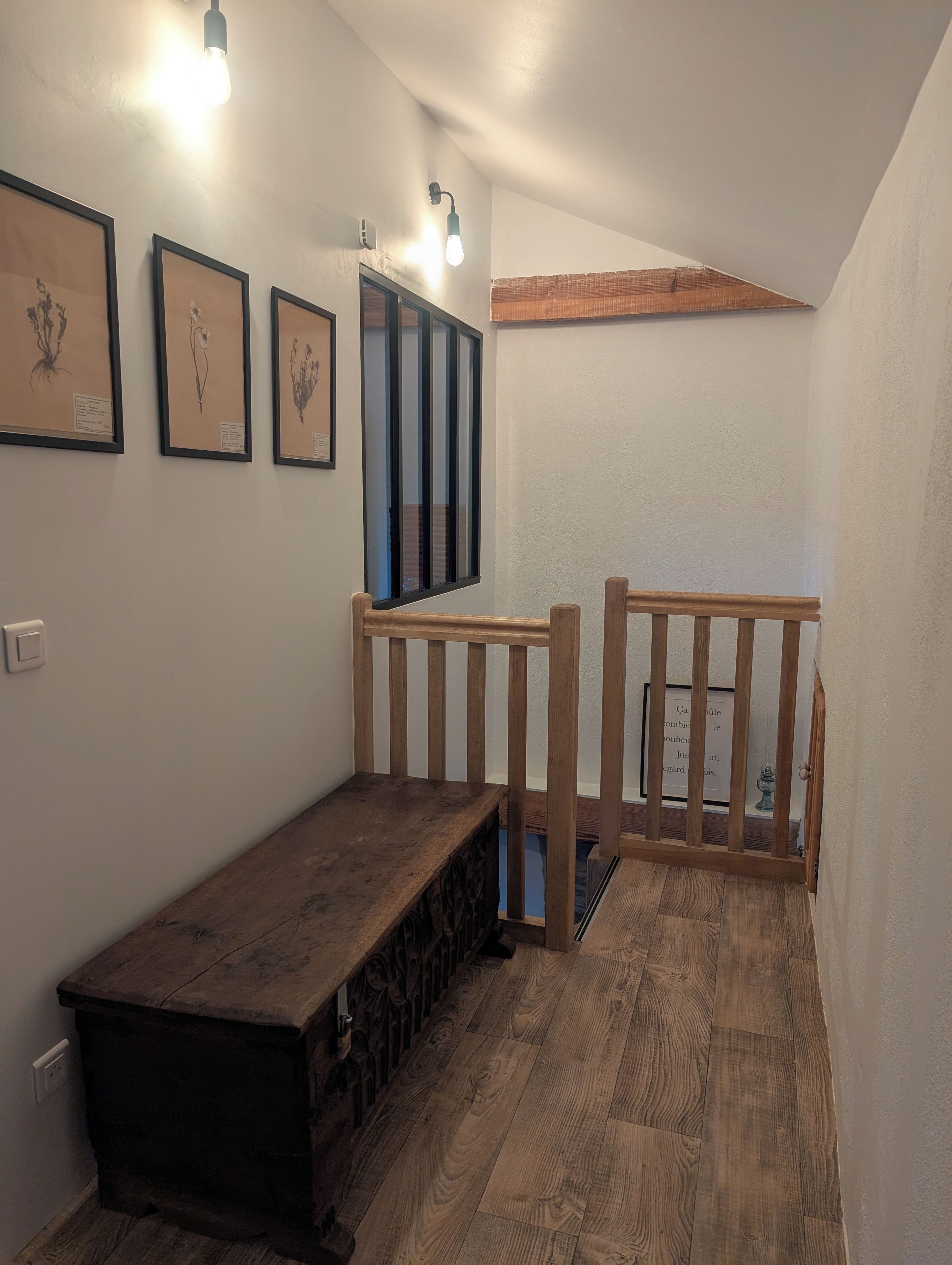Een kleine kamer met een houten bankje, een raam met verticale zonwering en ingelijste afbeeldingen van planten aan de muur.