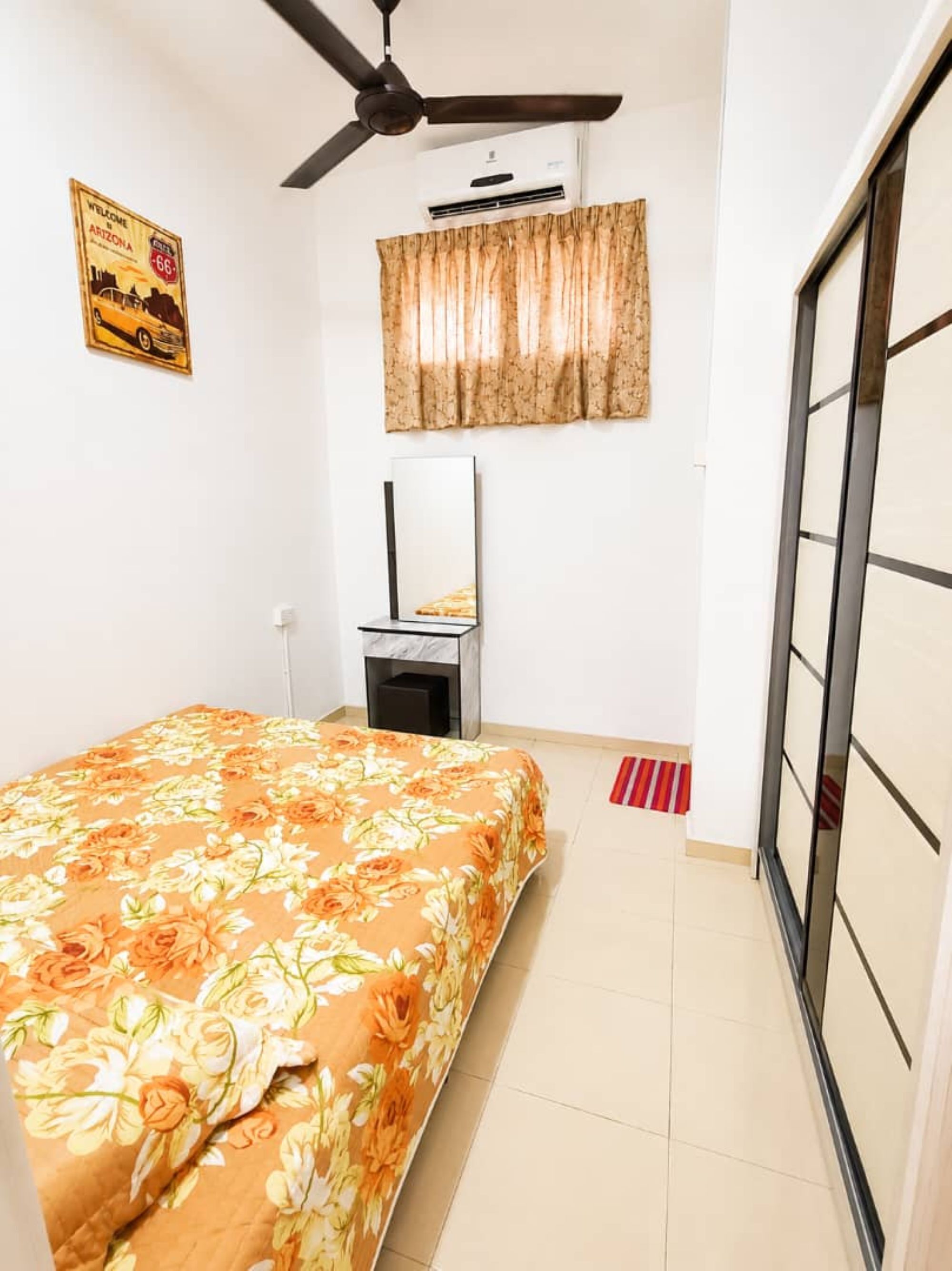 Una camera da letto con un letto, una ventola a soffitto, un condizionatore d'aria, una finestra con tende, uno specchio e una porta con pannello in vetro scorrevole.