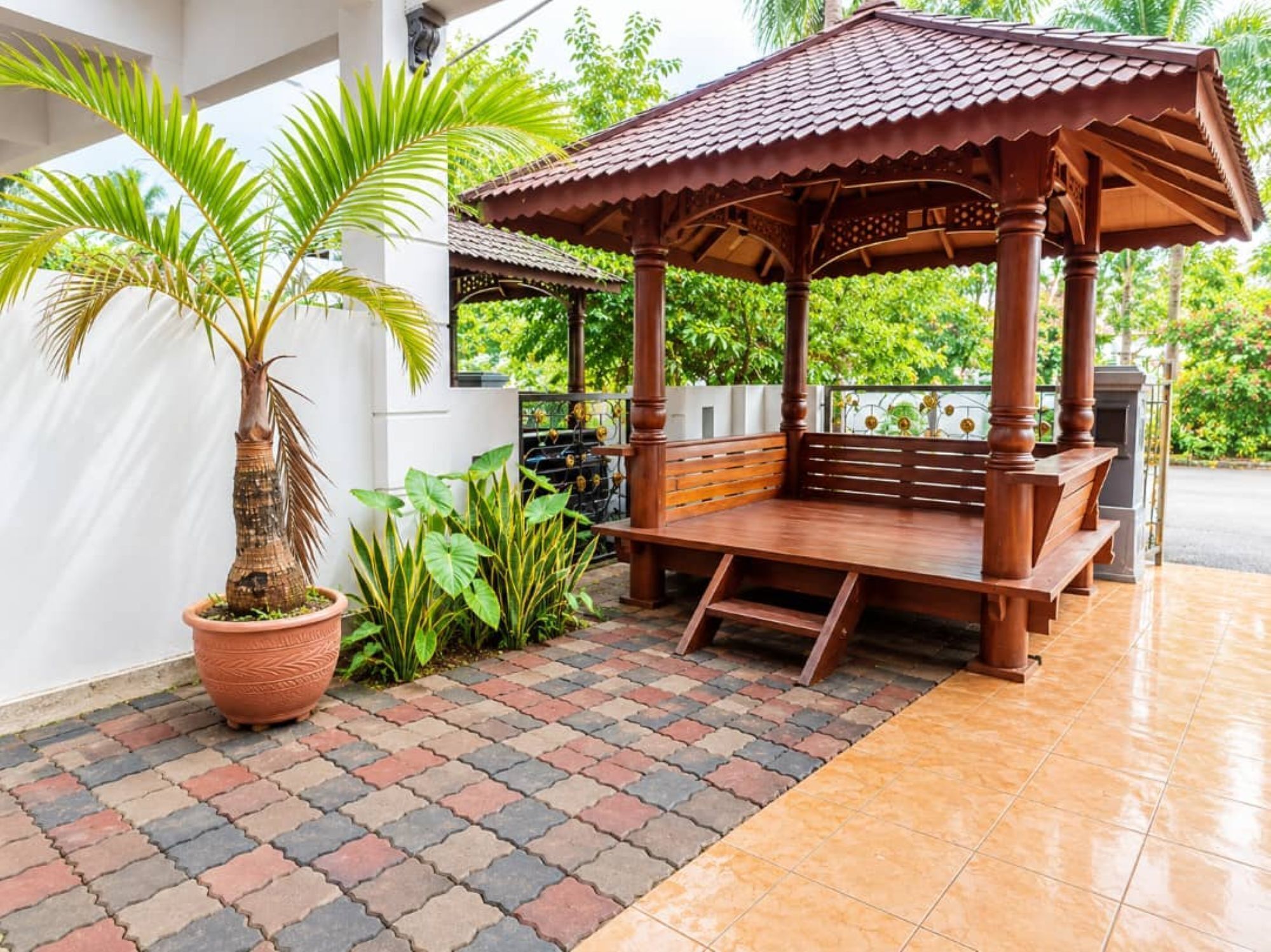 A wooden gazebo with a bench, surrounded by potted plants and a tiled patio.