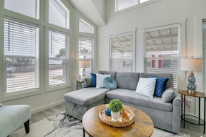 A modern living room with a grey sectional sofa, a round wooden coffee table, and a view through large windows with blinds. - 5 Mi to Beach: Family Fun, Modern with Great Views (Port Isabel)