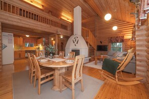 Interior de una cabaña de troncos con comedor, chimenea, sofá y mecedora.