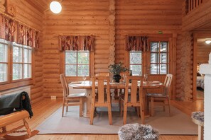 A wooden cabin interior with a dining table set for four, surrounded by chairs, and a fireplace visible in the background.