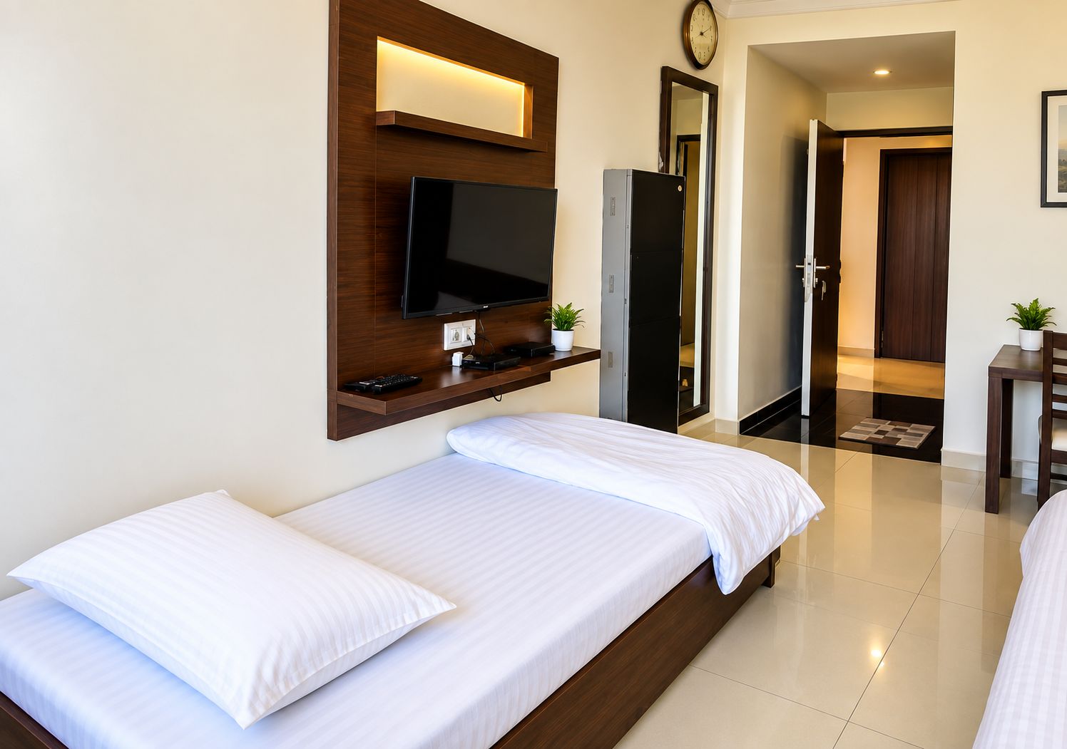 Habitación de hotel con dos camas individuales, un televisor montado, un reloj y una mesita con una planta.