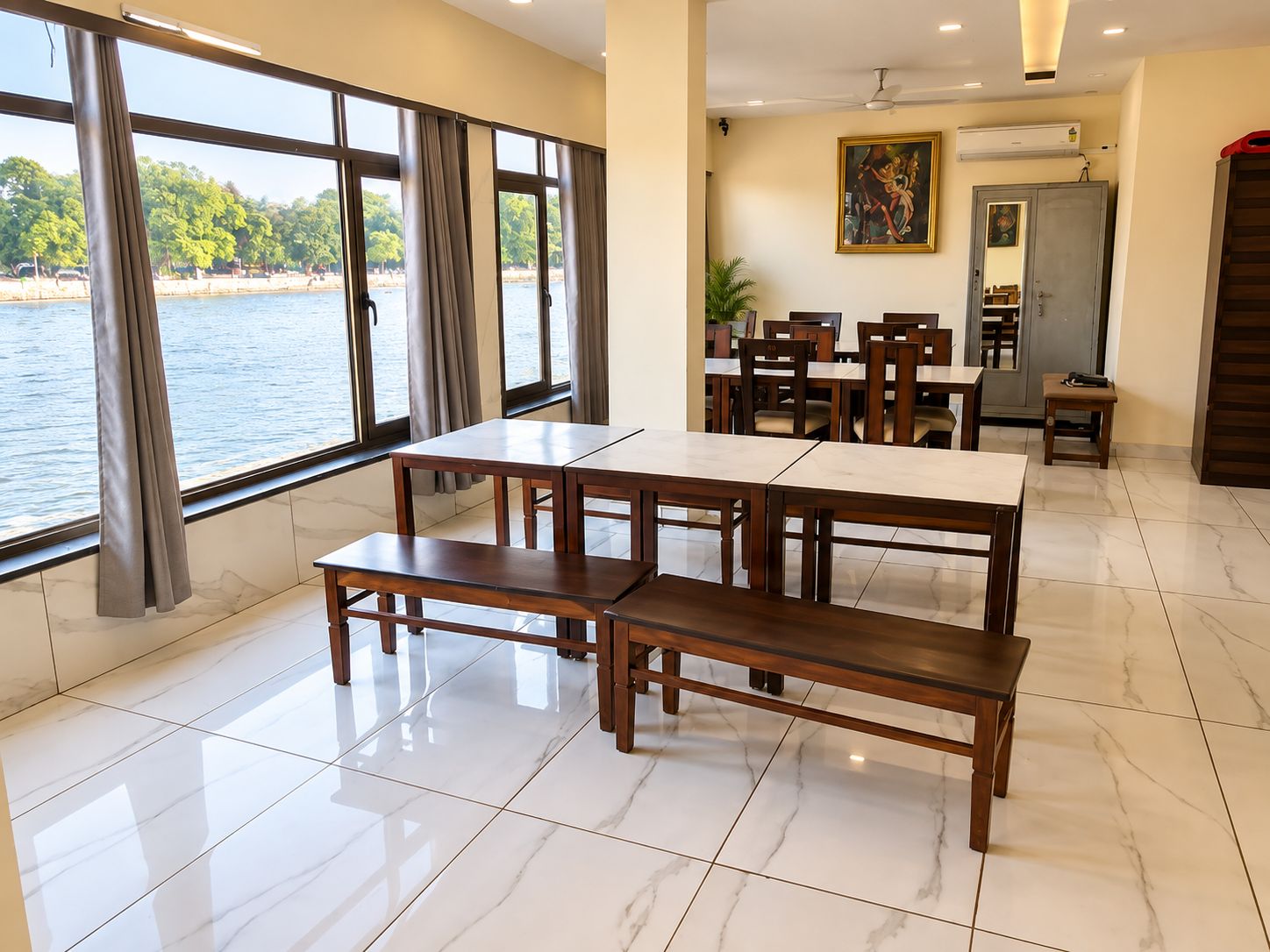 A dining area with a view of a body of water, featuring a table and benches, a ceiling fan, and a painting on the wall.