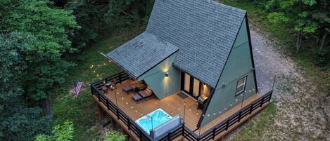 A cabin with a hot tub, deck, and outdoor seating area surrounded by trees.