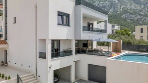 A modern multi-story building with a pool, outdoor seating, and a mountain backdrop. - Villa Baculla by Villas Guide (Makarska)