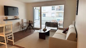 Ein Wohnzimmer mit einer Couch, einem Couchtisch, einem an der Wand befestigten Fernseher, einem Esstisch mit Stühlen und Blick auf den Balkon.