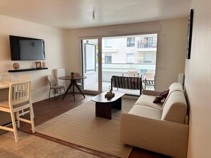A living room with a sofa, a coffee table, a TV mounted on the wall, a dining table with chairs, and a view of the outside balcony. - Charming Colombes Apartment  Comfort, Convenience, and Style (Colombes)