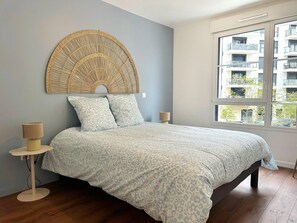 A bedroom with a bed, a wall-mounted headboard, a bedside table with a lamp, and a window with a view of buildings. - Charming Colombes Apartment  Comfort, Convenience, and Style (Colombes)