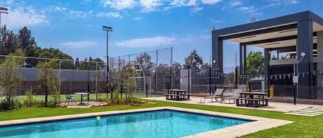 A swimming pool with a surrounding deck, a covered seating area, and a fenced sports court.