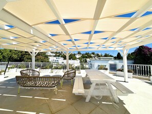 An outdoor patio with white furniture, a pergola with blue-tinted panels, and a view of white buildings in the background. - Appartement Rayhana Sidi Bou Said (Sidi Bou Said)