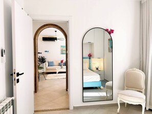 Camera d'albergo con un letto, una sedia, uno specchio e vista su un'altra stanza con un divano e una pianta.