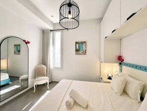 Una camera da letto con un letto, una sedia, uno specchio, una finestra con tende e un quadro appeso al muro.