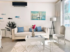 Un salon avec un canapé beige, une table basse blanche, un lampadaire et un tableau représentant des voiliers.