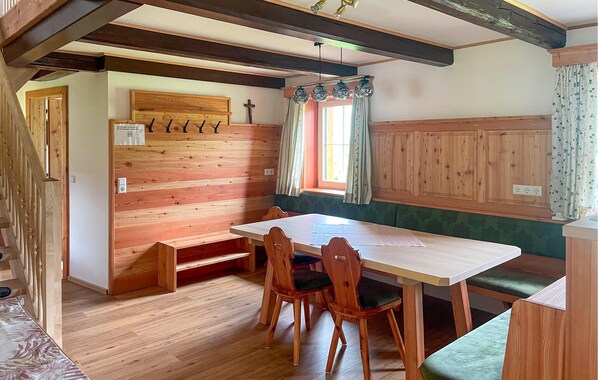 A wooden interior with a table and chairs, a bench, a window with curtains, and a cross on the wall.