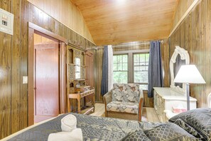Ein Zimmer mit Holzwänden, einem Bett, einem Sessel, einem Schminktisch und einem Fenster mit Vorhängen.