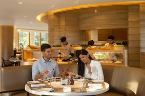 Ein Restaurant mit einem runden Tisch für zwei Personen, einem Koch, der im Hintergrund kocht, und Blick ins Freie durch große Fenster.