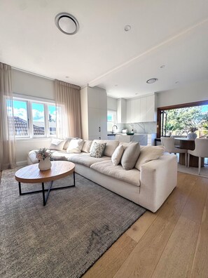 Una sala moderna con un sofá seccional beige, una mesa redonda de centro y vista a la cocina a través de una puerta abierta.
