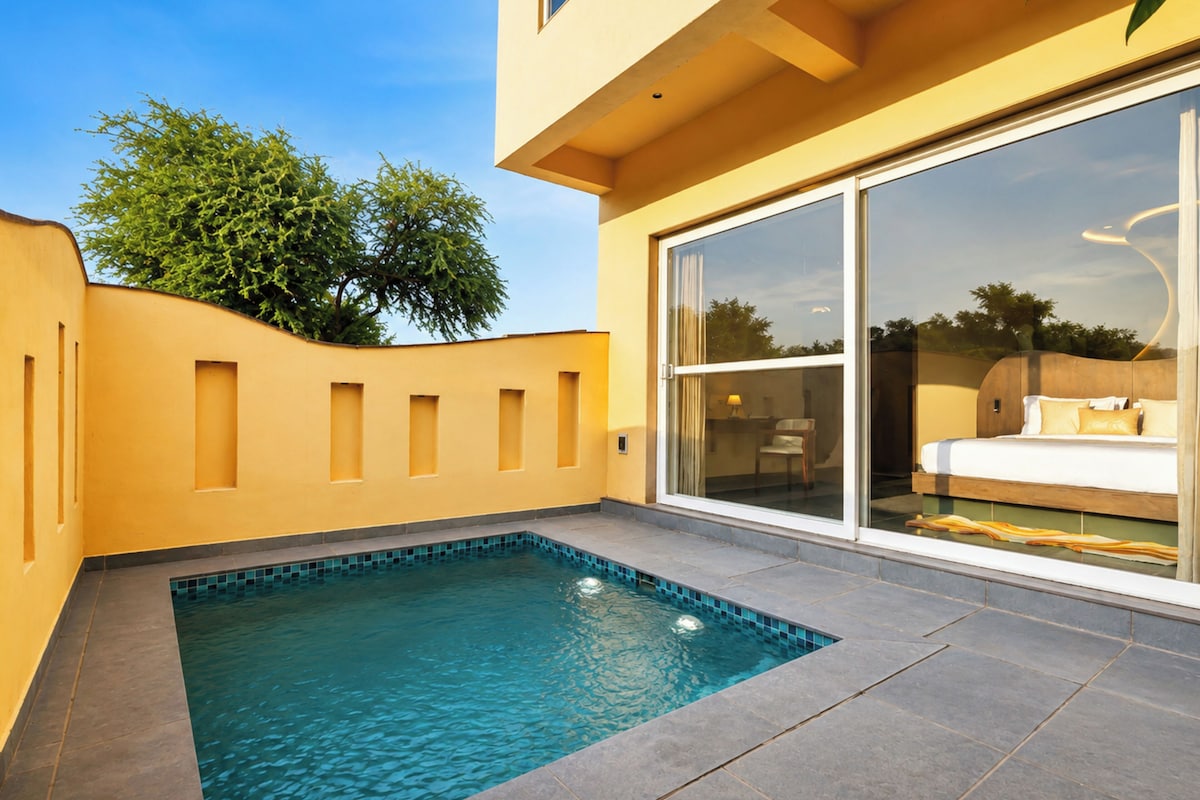 A modern pool area with a clear blue pool, a yellow wall, and a large sliding glass door leading to a bedroom with a bed and a chair.