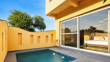 Un área de piscina moderna con una piscina azul cristalina, una pared amarilla y un amplio ventanal corredera que da a un dormitorio con una cama y una silla.