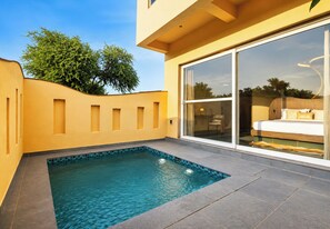 Un área de piscina moderna con una piscina azul cristalina, una pared amarilla y un amplio ventanal corredera que da a un dormitorio con una cama y una silla.