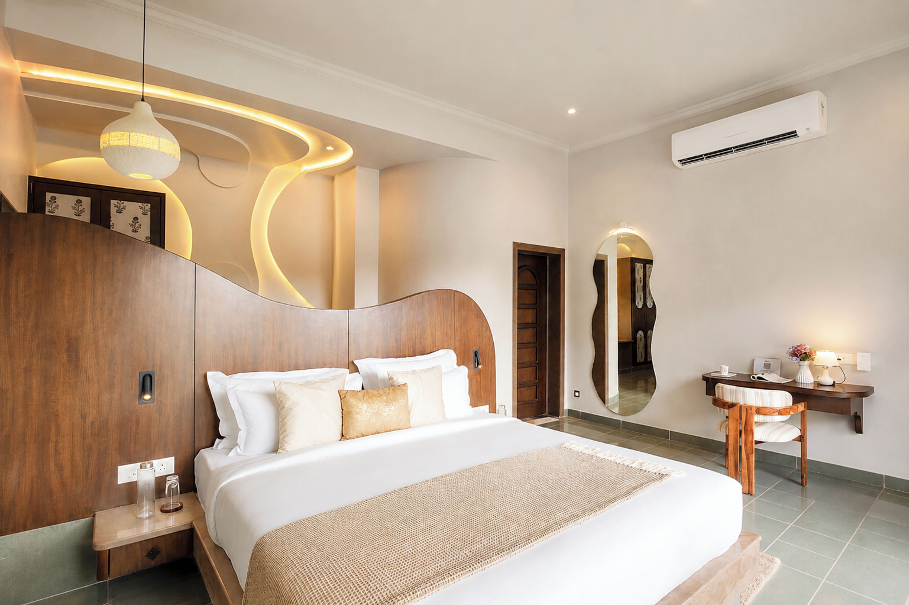 A modern bedroom with a large bed, a wooden headboard, a bedside table, a nightstand, a mirror, a desk with a chair, and an air conditioning unit.
