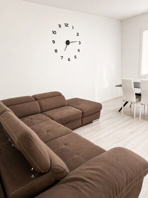 Ein modernes Wohnzimmer mit einem Ecksofa, einer Wanduhr und einem Essbereich mit Stühlen.