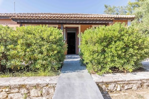 A building with a tiled roof, a stone pathway leading to the entrance, and well-maintained green shrubs on either side. - Zanteva-Psophis cottage (Zakynthos)