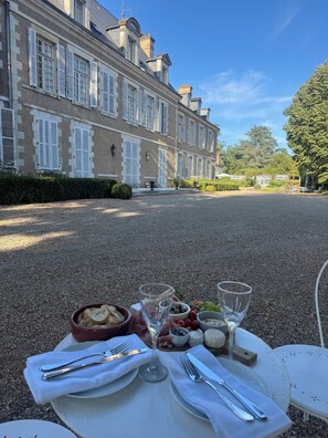 Montaje de comedor al aire libre con una mesa dispuesta para dos personas, mostrando una variedad de comida y bebidas, frente a un edificio grande con múltiples ventanas y un jardín cuidado.