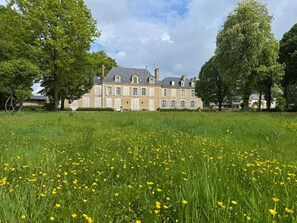 A large, historic building with multiple windows and a symmetrical facade, surrounded by a well-maintained lawn with yellow flowers.