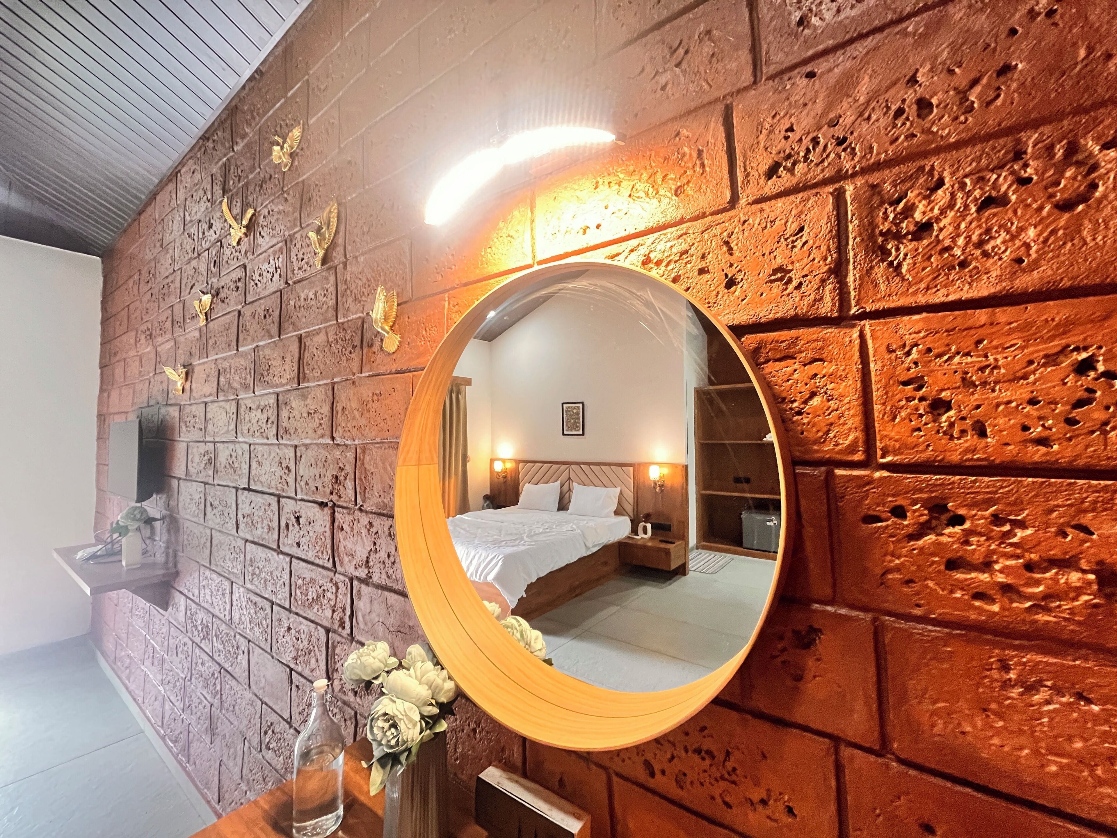A bedroom with a brick wall, a round mirror, a bed, bedside tables, and a wall-mounted TV.