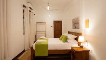 Habitación de hotel con cama, mesita de noche, lámpara, ventilador de techo y un cuadro en la pared.