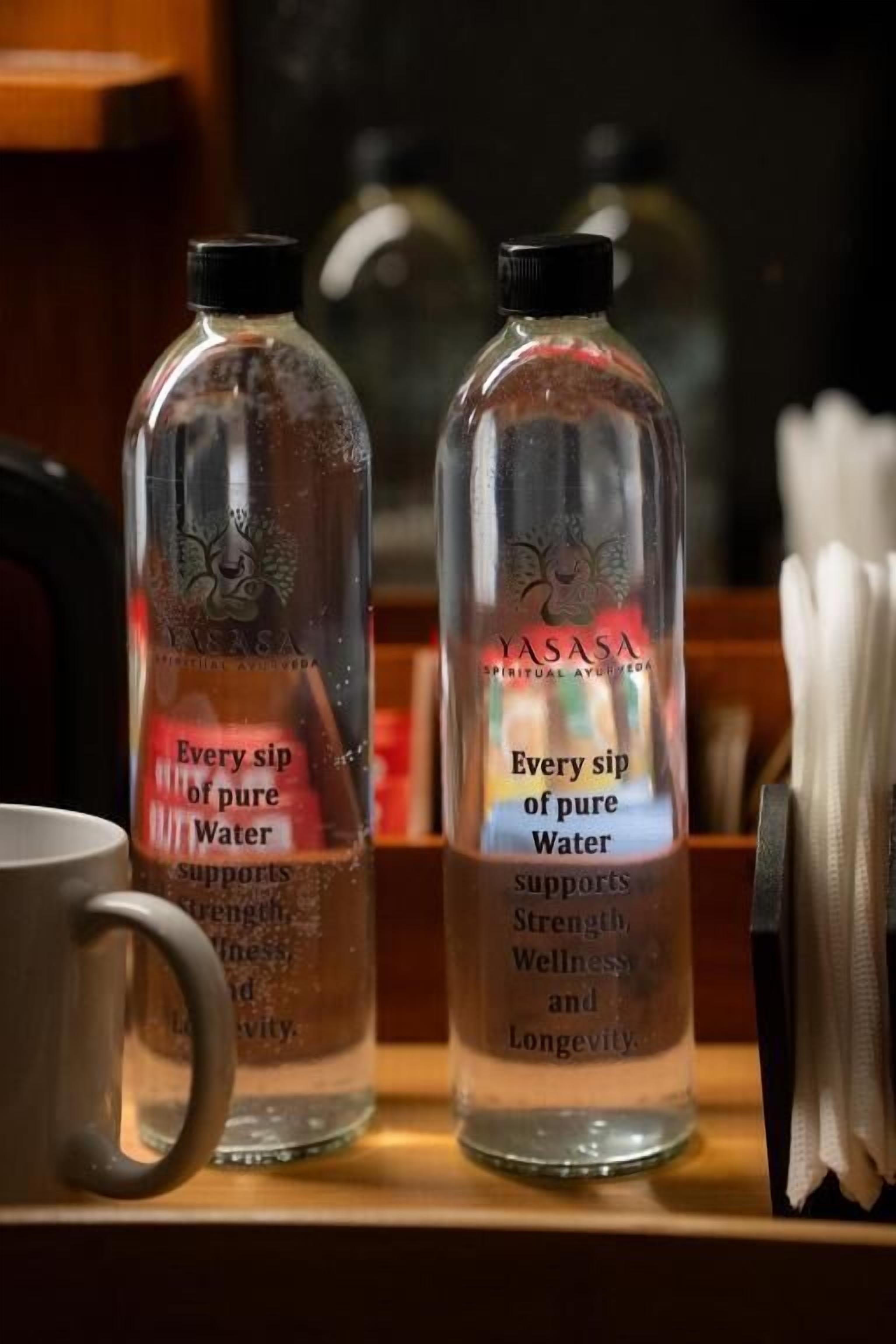 Dos botellas de vidrio transparente etiquetadas 'YASASA' con texto que promociona beneficios para la salud.